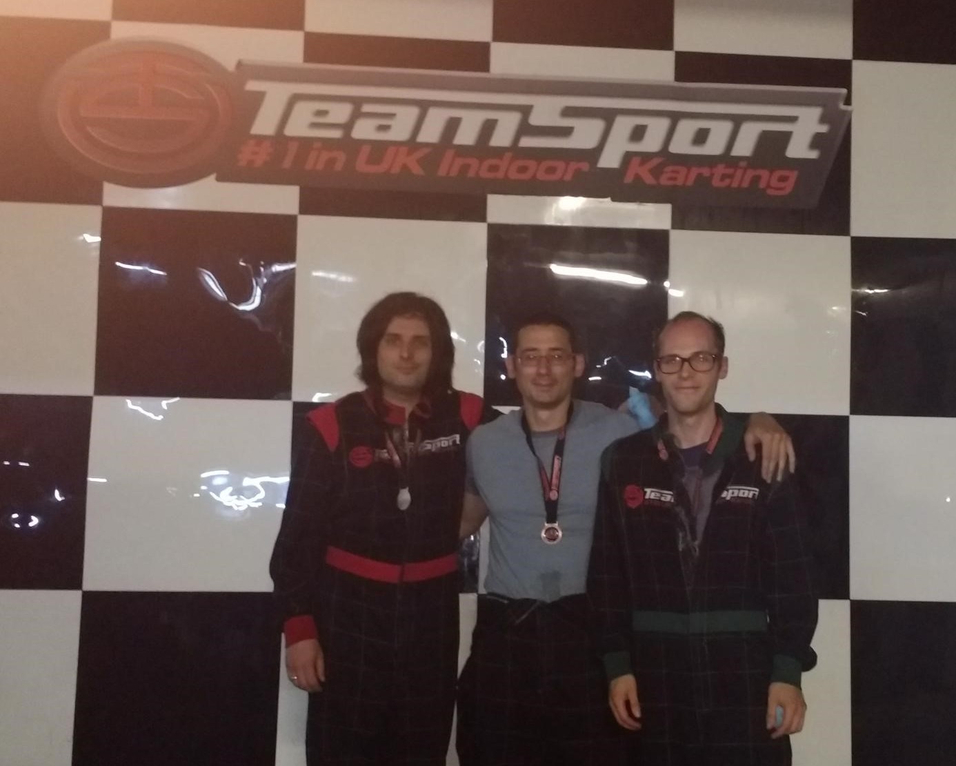 Three people in karting attire arm to arm standing in front of a sign that saus Team Sport #1 in UK Indoor Karting